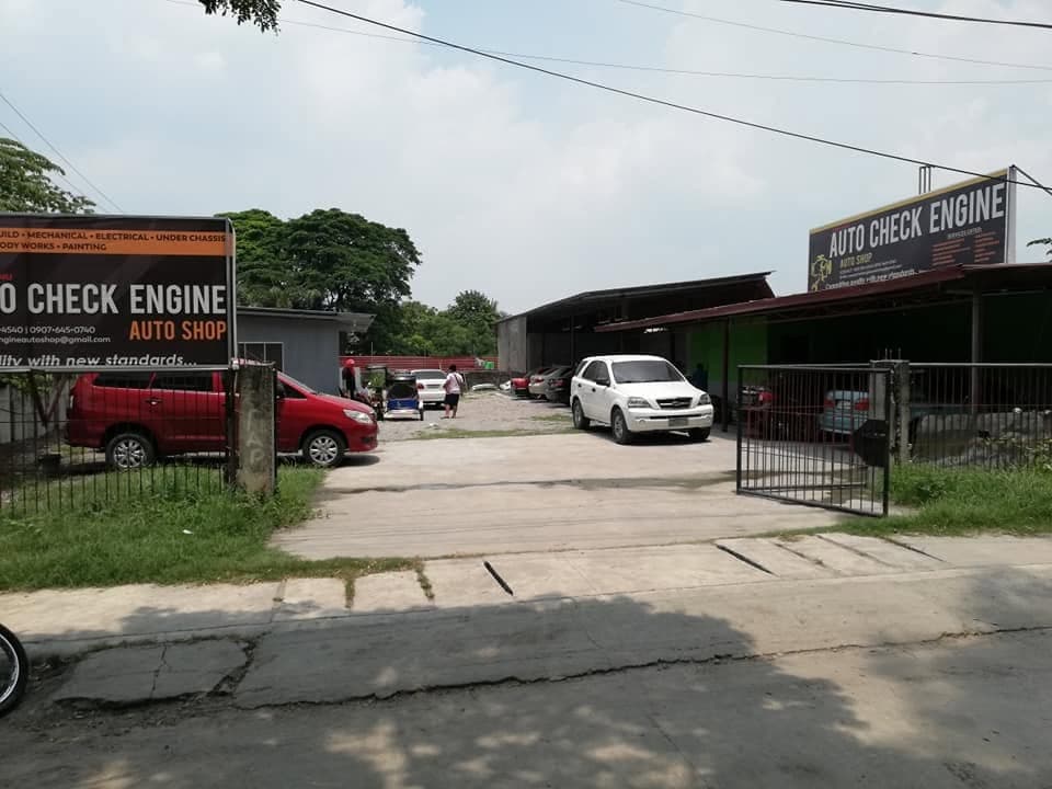 The entrance to Auto Check Engine from the road, with signs on both sides of the gate and vehicles parked in the yard
