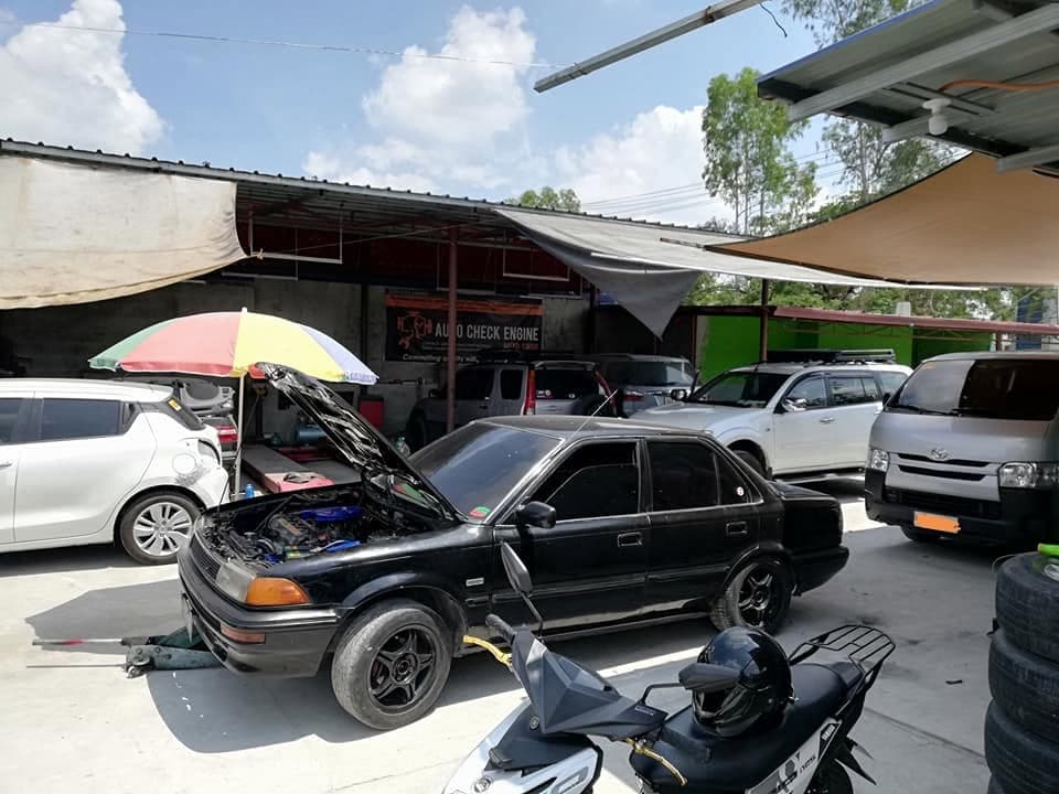 An older black Toyota Corolla with its bonnet up under a parasol, other vehicles waiting behind it