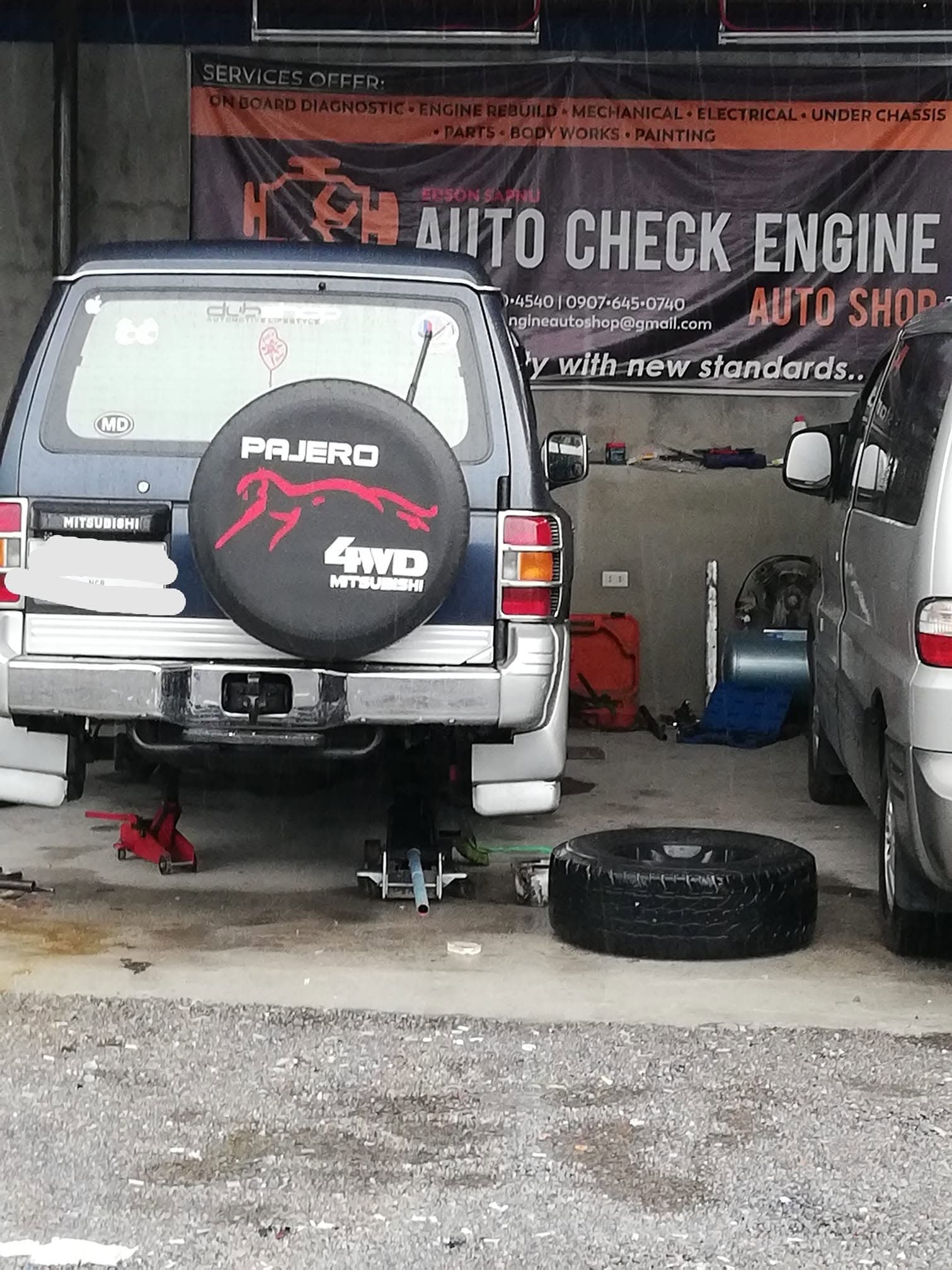 A Mitsubishi Pajero raised on a trolley jack and axle stand with a rear wheel removed, the wheel lying on the floor beside it