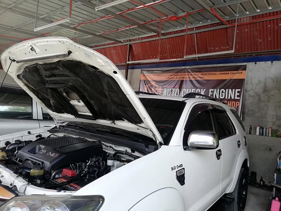 A white Toyota Fortuner 3.0 D-4D in the covered bay with its bonnet raised, the shop banner on the wall behind