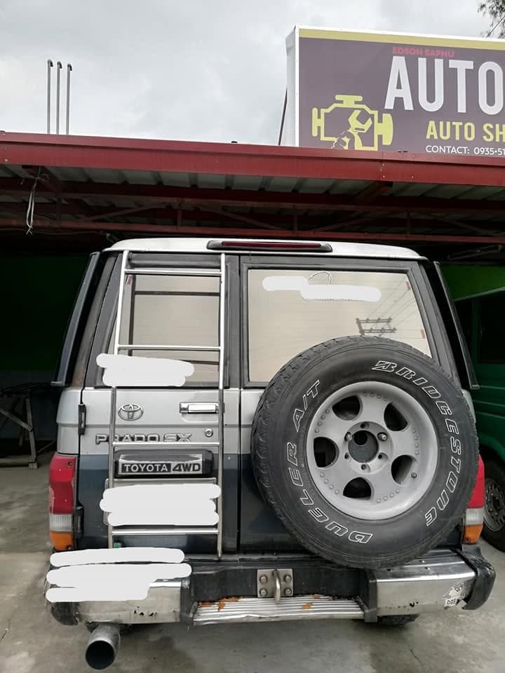 The back of a Toyota Land Cruiser Prado SX with its spare wheel mounted on the tailgate, parked under the shop sign